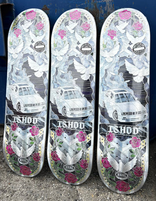 Three skateboard decks standing upright side-by-side, featuring a graphic design with white cars, white doves, pink roses, and green leaves on a white background, with the brand name 'ISHOD' printed on the bottom.