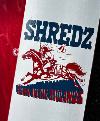 A close-up of a white skateboard deck featuring a red graphic of a cowboy riding a galloping horse and the text 'SHREDZ' and 'BORN IN THE BADLANDS' in red and navy blue.
