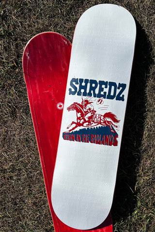 Two skateboard decks laid flat on grass, with one white deck featuring a red and blue 'SHREDZ' graphic partially covering a red deck underneath.